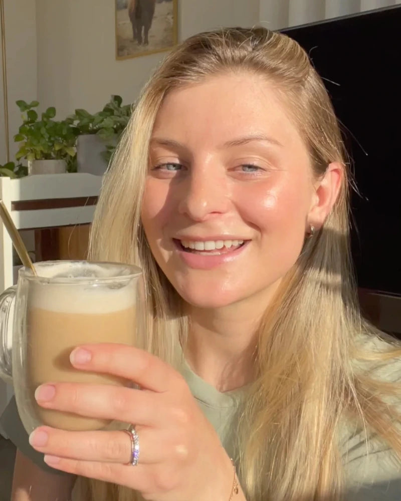 A person with long blonde hair smiles while holding a glass mug filled with a frothy beverage. Sunlight creates a warm glow on their face. A chair, plants, and a framed picture are visible in the bright background.